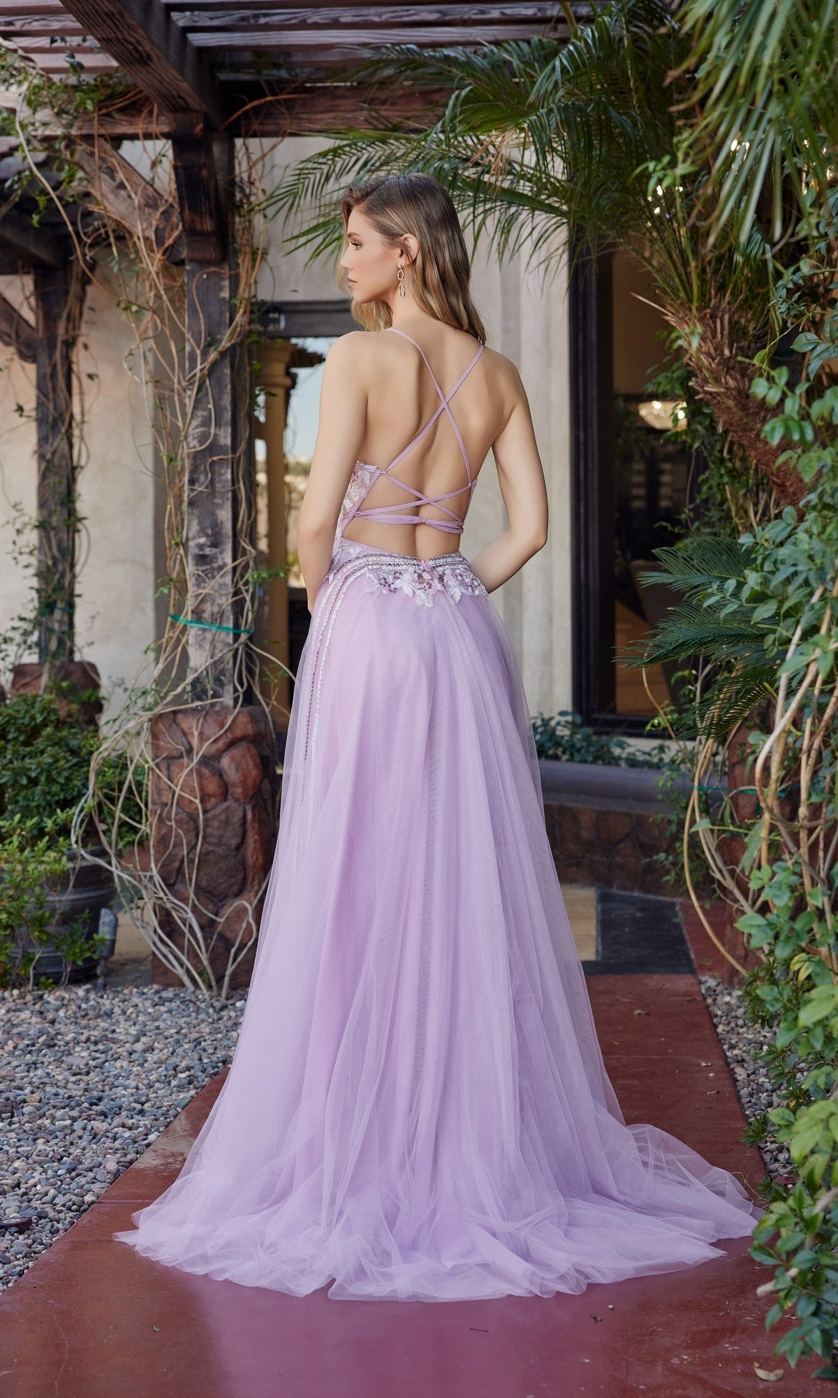 A woman stands outdoors in the Nox Anabel G1795, a light purple long prom dress featuring an open back, floral waist embellishments, and an elegant a-line tulle skirt, surrounded by lush greenery.
